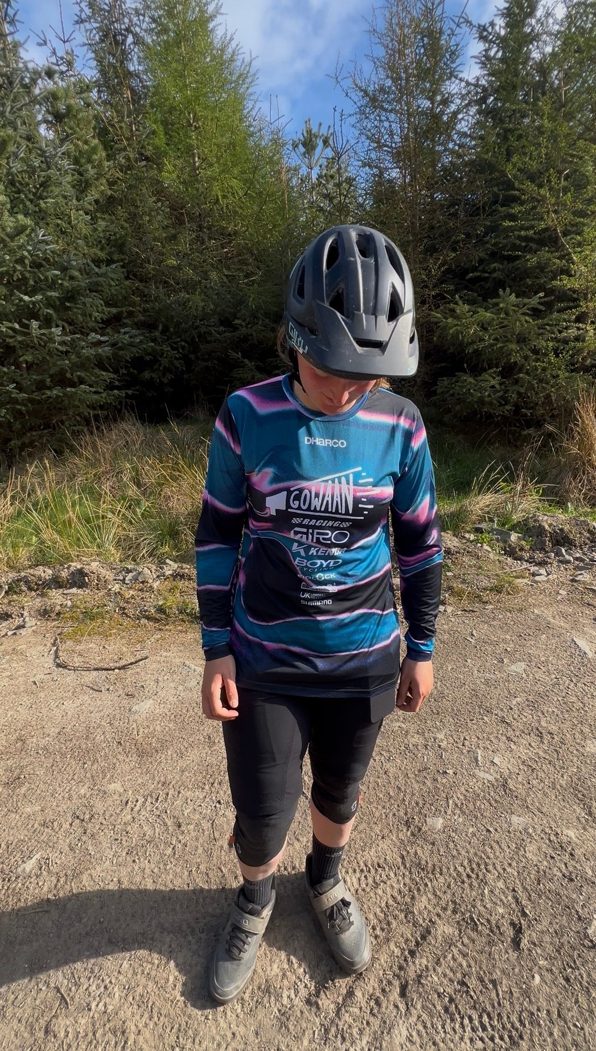 Person wearing the colorful Gowaan Racing Dharco cycling jersey and helmet on a dirt path with trees in the background